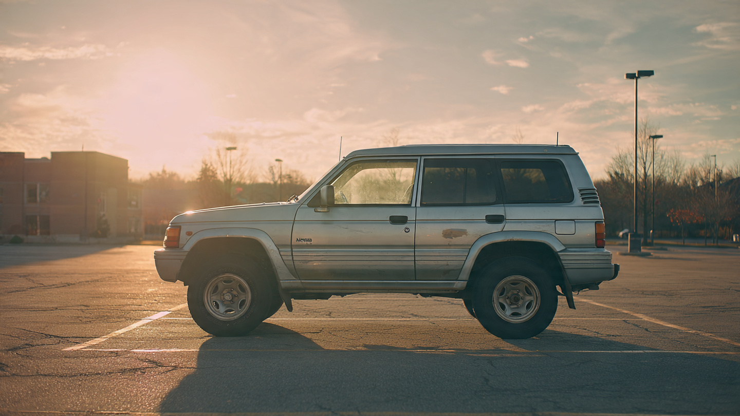 An aging Mitsubishi Montero SUV parked in a dusty lot, sun-bleached paint and worn tires